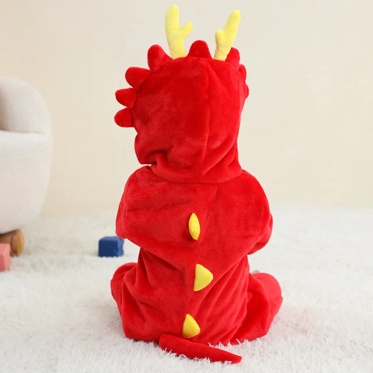 Et billede af et sejt rødt dinosaurkostume til børn, udført i blødt stof med en gul mave. Dragten har en detaljeret hætte med et dinosauransigt og hvide takker, der fortsætter ned ad ryggen til den lange hale.