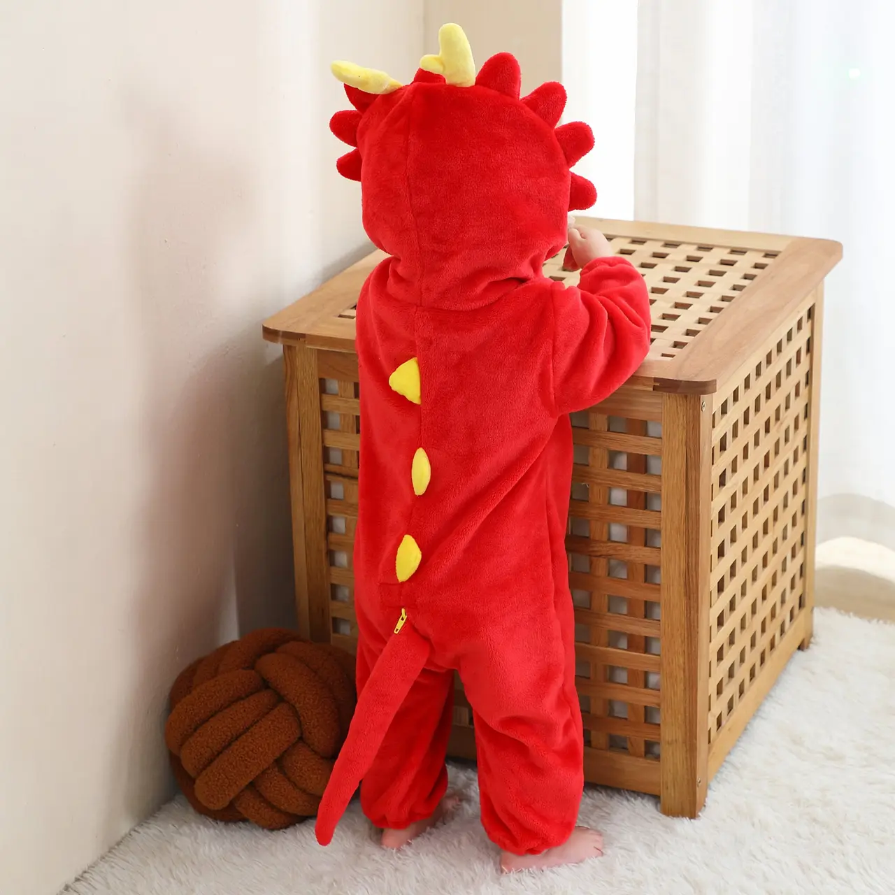 Dette billede viser et nuttet og blødt rødt dinosaurkostume til børn, set bagfra for at fremhæve de takkede pigge langs ryggen og den lille hale. Dragten er lavet af behageligt flannelmateriale og har en matchende hætte med øjne og tænder, hvilket gør den perfekt til leg eller fastelavn.