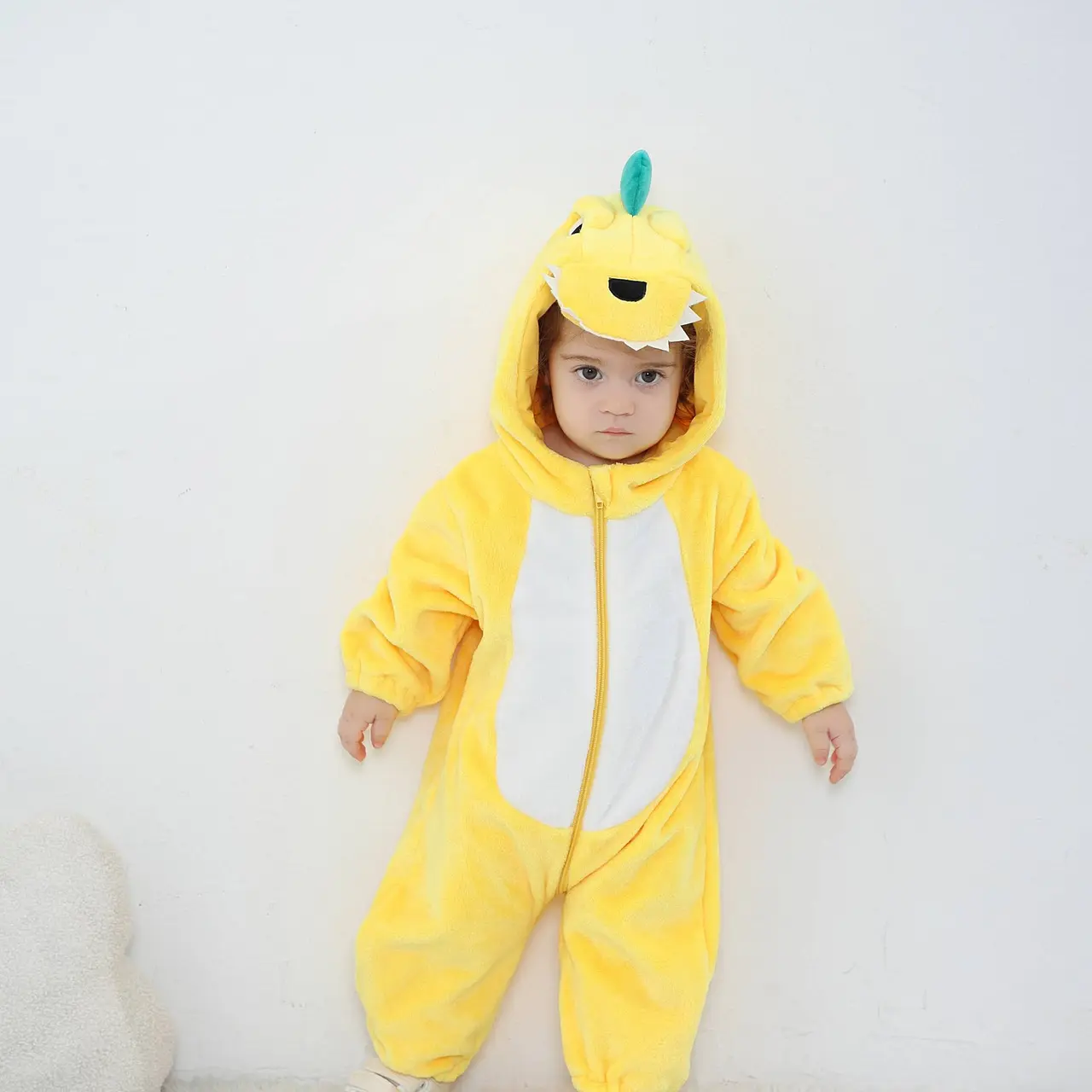 Et charmerende og blødt gult dinosaur-kostume til børn, formet som en heldragt i behageligt flannelmateriale. Dragten har en hvid mave, en række orange pigge ned langs ryggen og en sød hætte med store øjne og små tænder, der fuldender det legesyge look.